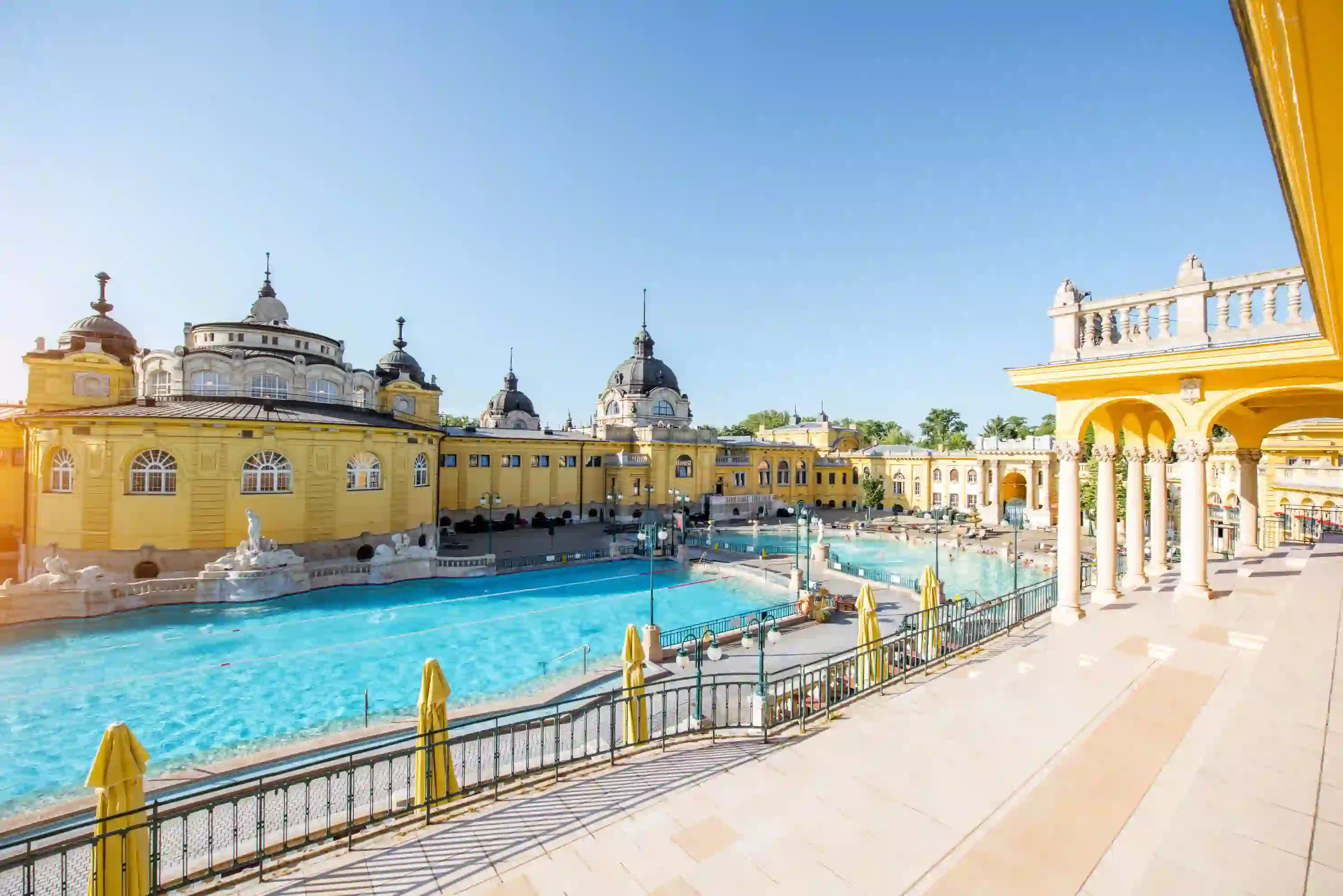 Szechenyi Thermal Baths, showing one large swimming pool and a smaller one to the right. Behind, a yellow building with grey domes and turrets on top.