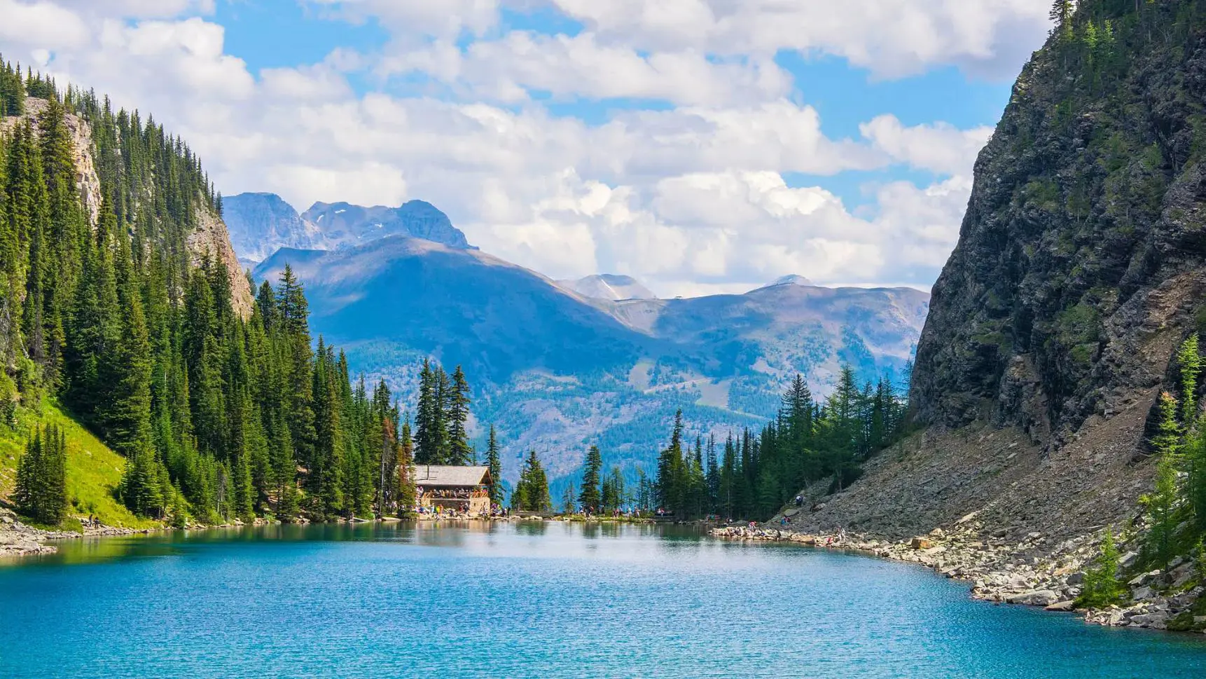 A clear turquoise lake with gentle ripples in Banff National Park, Alberta, Canada, bordered by lush green pine forests and rolling hills, with a small cabin situated at the far end of the lake