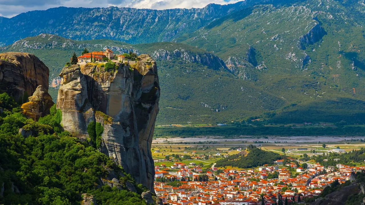 High rock precipice with a monastry on the top. In the background, mountains and a town on the land below 