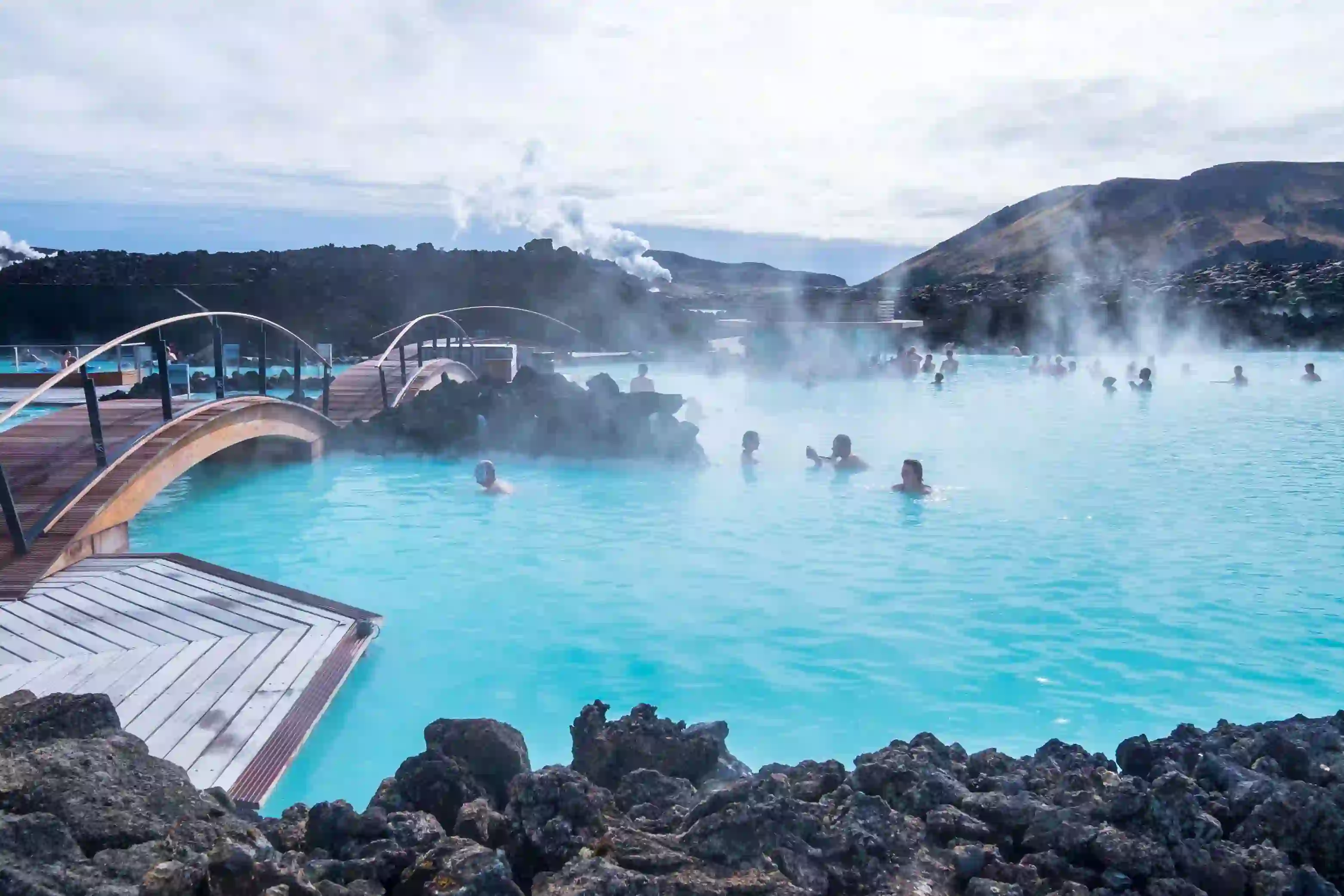 Geothermal lagoon in Iceland - bright blue water with steam coming off, surrounded by black rock. People swimming.