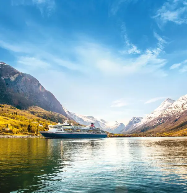 The Balmoral cruise ship sails through a tranquil fjord in Norway, surrounded by steep, snow-capped mountains and lush, forested hills beneath a bright blue sky