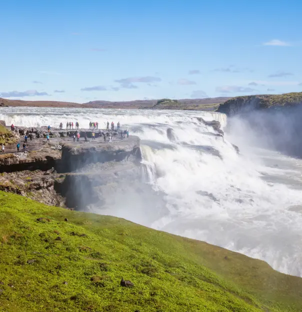 High angle shot of the mouth of a waterfall. People stood on the rock next to it, grassy land in the forefront