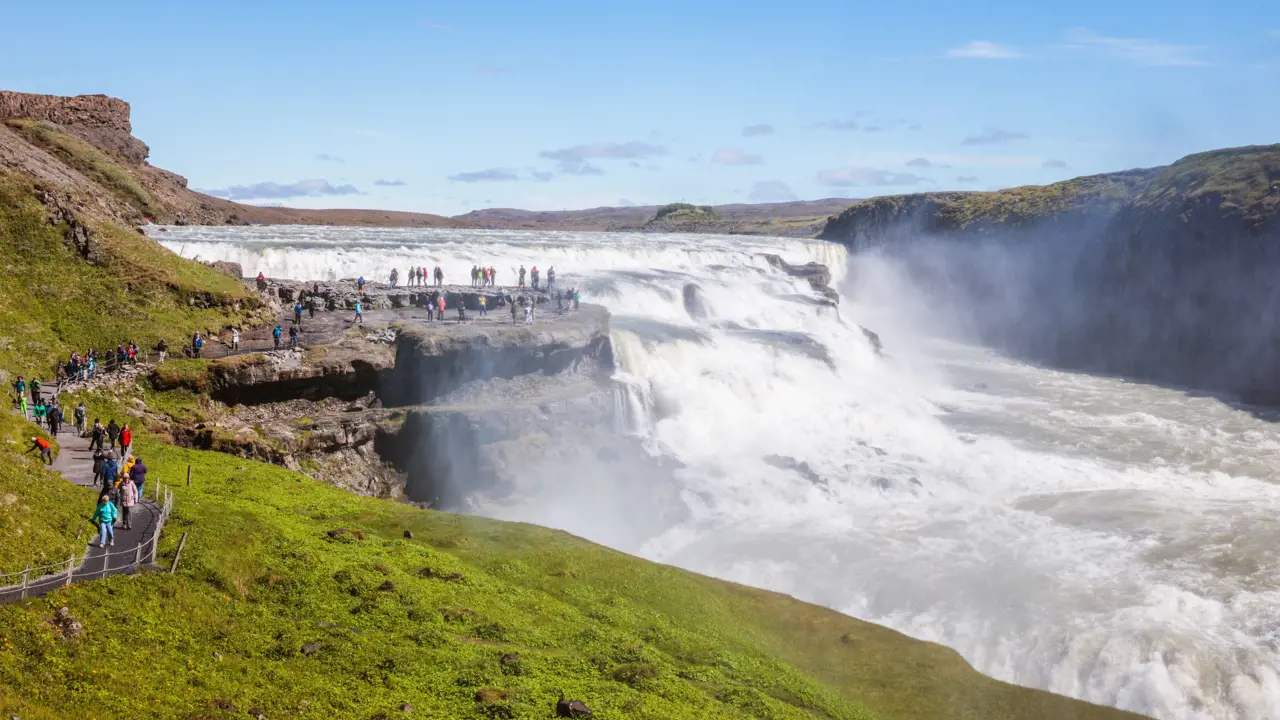 High angle shot of the mouth of a waterfall. People stood on the rock next to it, grassy land in the forefront