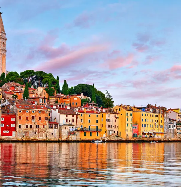 View of the town of Rovinj from the water, with colourful stone buildings on the waterfront, rows of these and trees going up the land behind and a tower with a statue on top. The sky is blue with pink clouds.