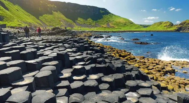 Large stones of the Giant's Causeway at the forefront, with waves crashing into them, back of people in the distance walking towards a grassy mountain