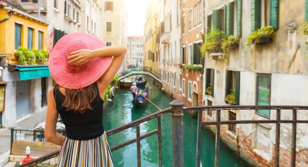 A woman in a striped skirt and pink hat standing on a bridge, looking out over a canal with gondolas in Venice, Italy, on a sunny day