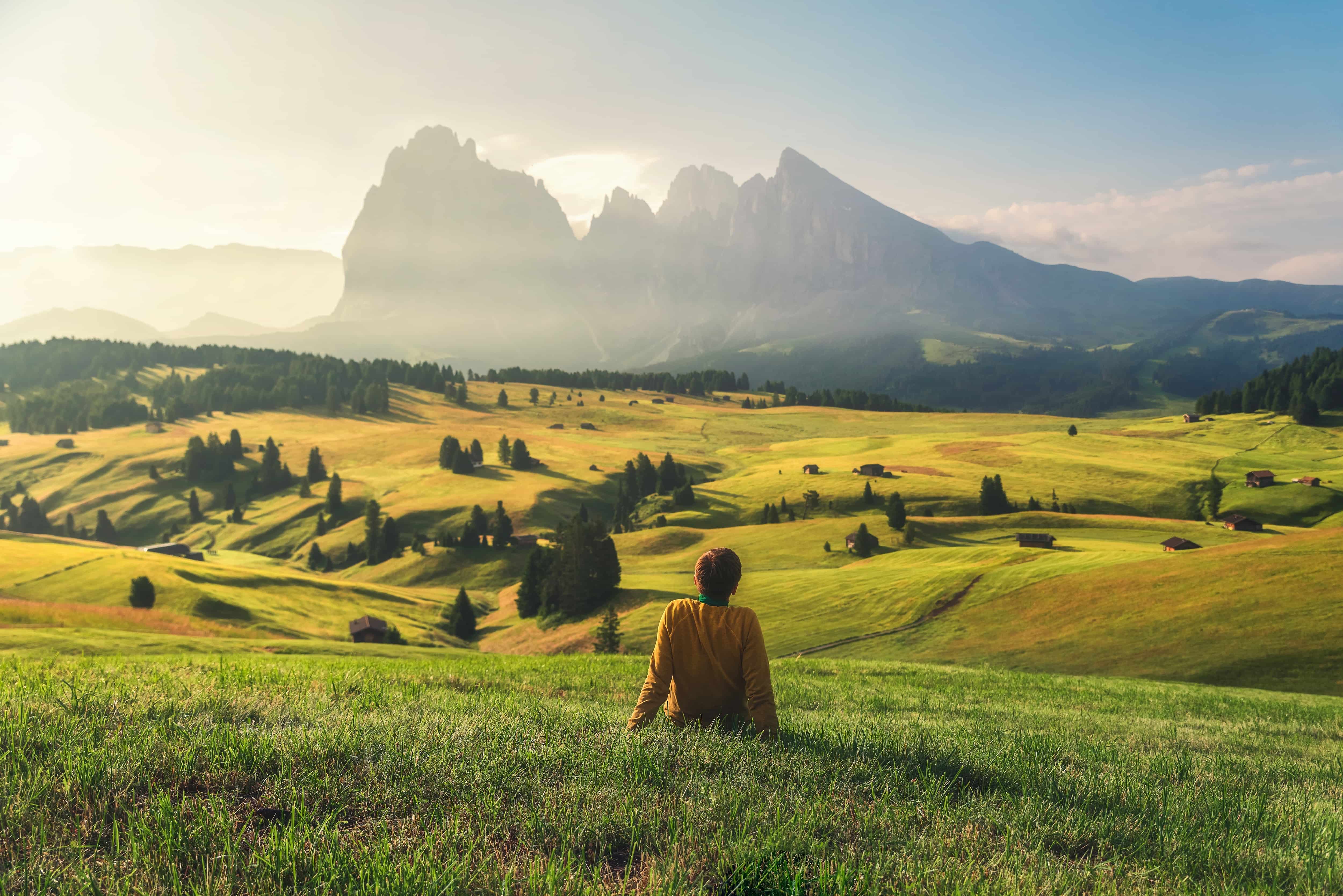 Shot of the back of a man sat looking out on the mountains of the Austrian Tyrol, covered in grass and fir trees dotted all over. In the distance, silhouettes of large mountains