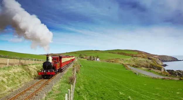 Red and black steam train heading towards the camera on a grassy hill on the coast of the Isle of Man, with a blue sky behind it