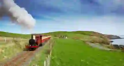 Red and black steam train heading towards the camera on a grassy hill on the coast of the Isle of Man, with a blue sky behind it