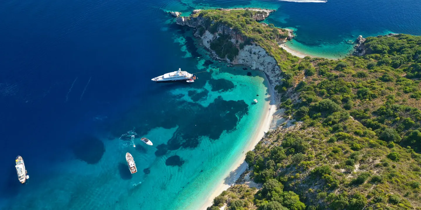 Bird's eye view of a sandy beach and bay of Gidaki, bright blue water with boats docked. Forested land in the right forefront