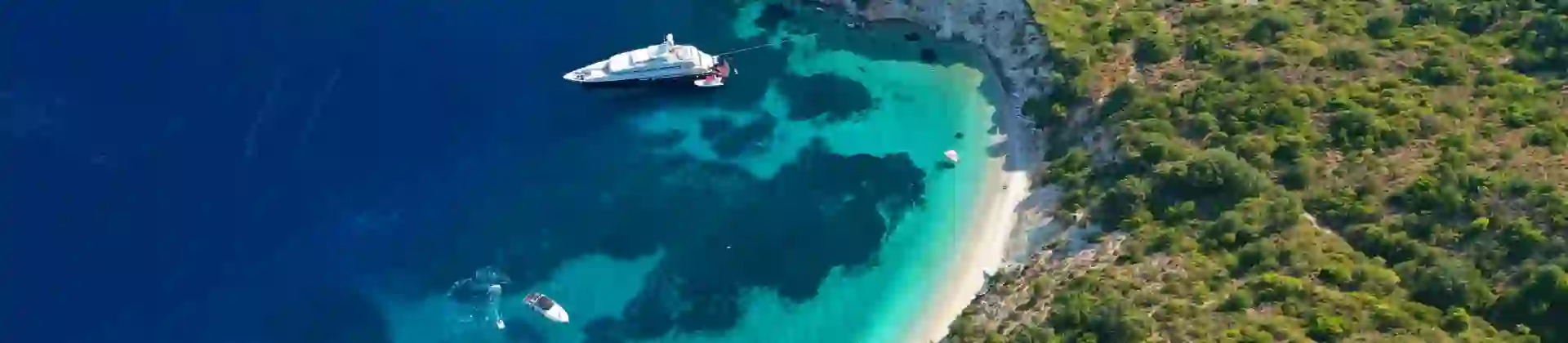 Bird's eye view of a sandy beach and bay of Gidaki, bright blue water with boats docked. Forested land in the right forefront
