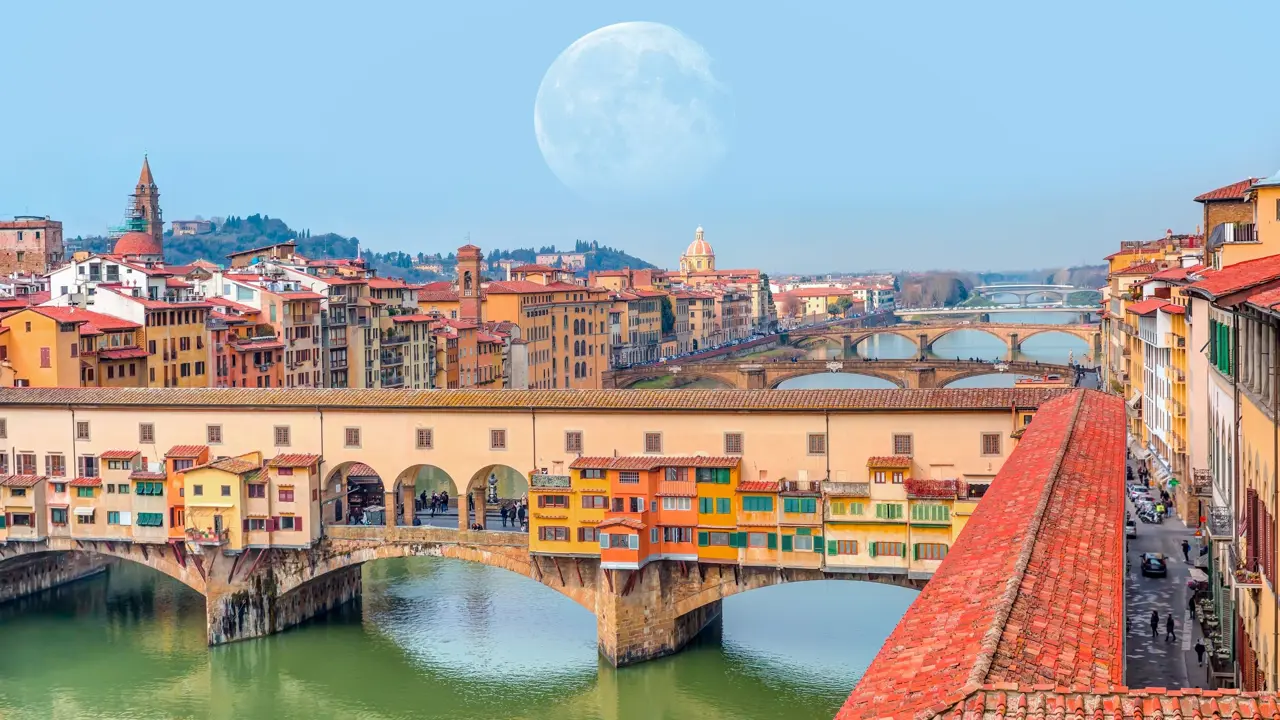 View of Ponte Vecchio Bridge showing buildings, the rest of The Arno River in the background, and a faint full moon in the sky