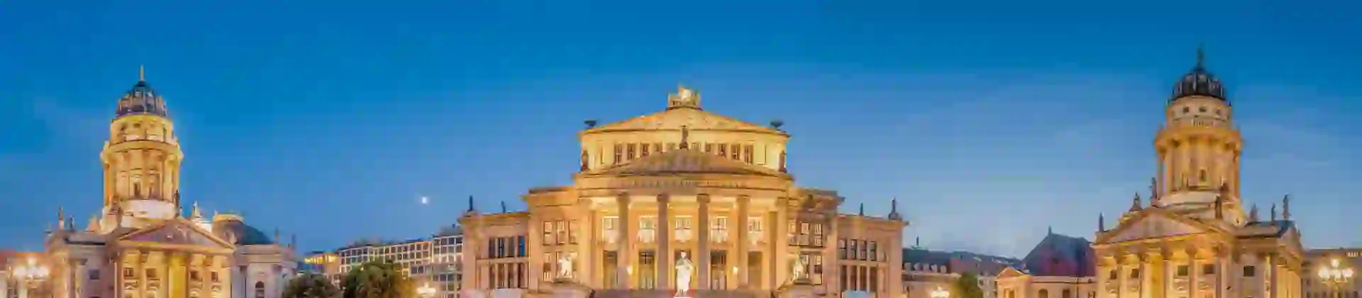 Wide angle shot of a grand, lit up building, with a building either side, both with a tower with a domed top. The middle building has a white statue of a person on a raised platform in front. The sky is a bright blue.