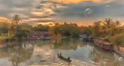 Two people in a canal boat on the Kerala Backwaters at sunset, with a pink house and palm trees lining the banks, and warm light reflecting on the calm water