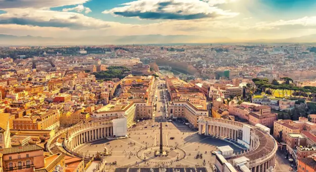 Aerial view of St. Peter’s Square in Vatican City, bathed in golden sunlight with Rome in the distance under a partly cloudy sky