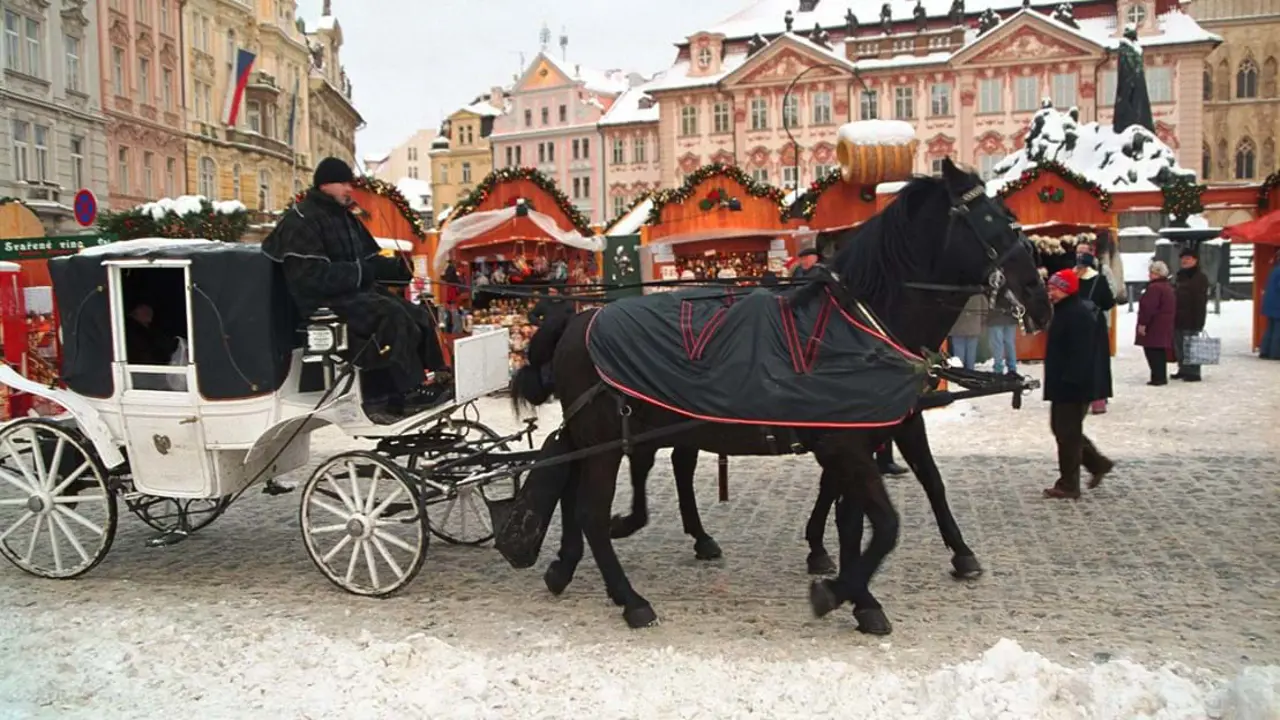 Black horses with black and red coats pulling a white cart with a man dressed in all back in it. Behind is some small market stalls with festive decorations, in front of some large buildings.