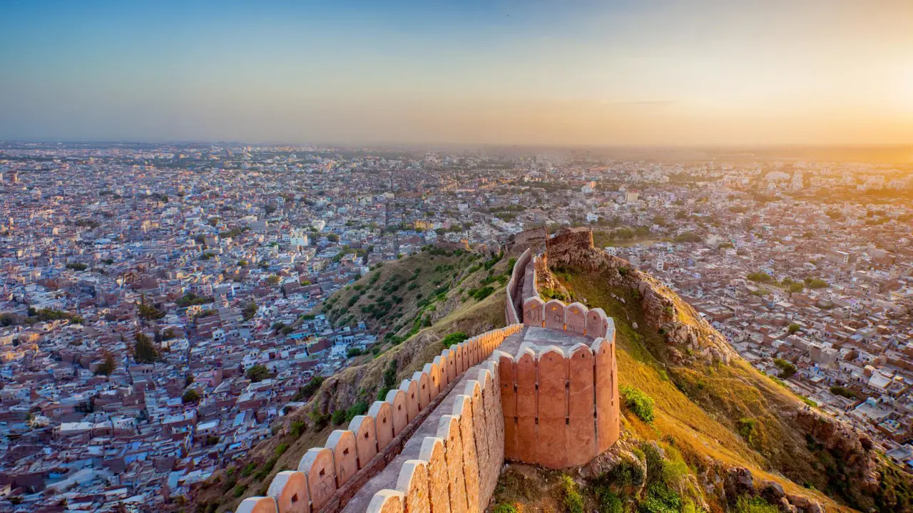 An aerial view of Nahargarh Fort in Jaipur stretching along a ridge at sunset, overlooking the sprawling cityscape below under a hazy orange sky