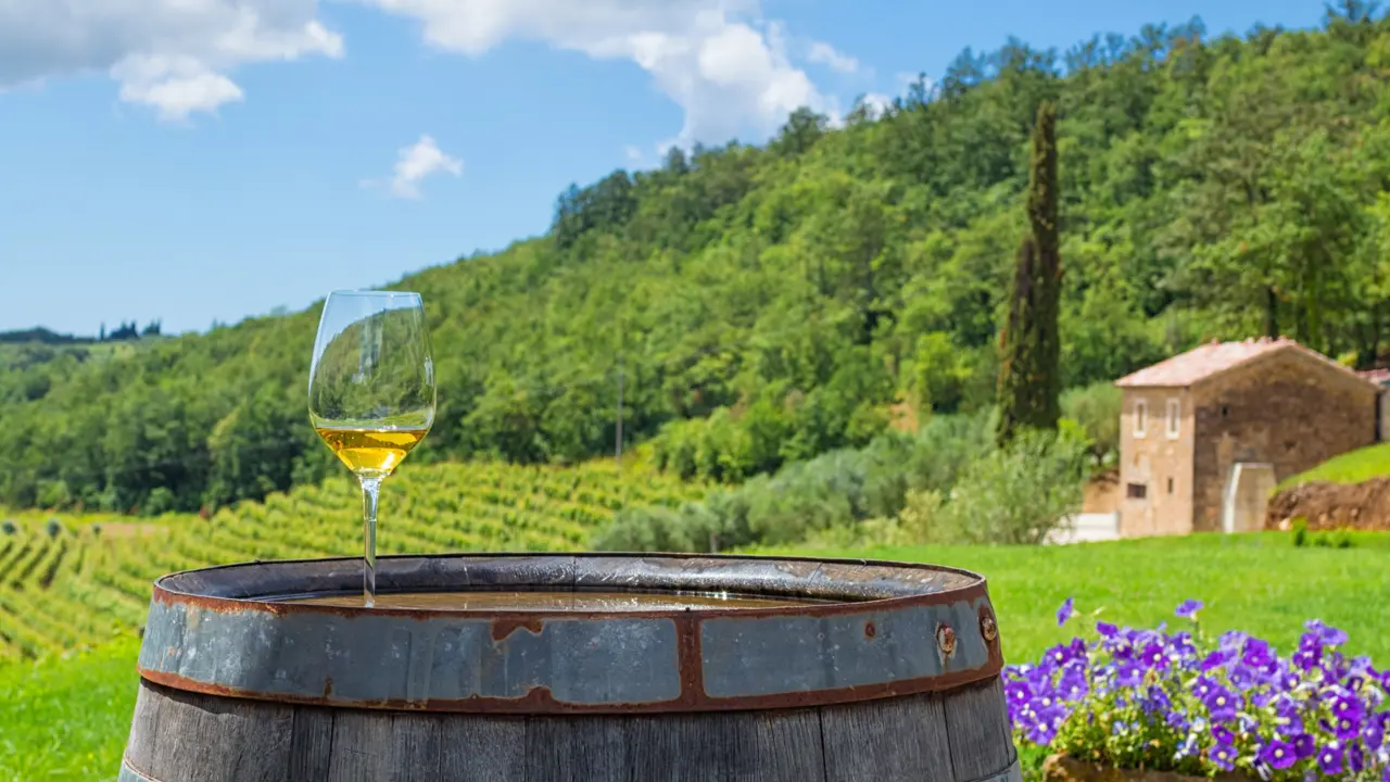 A glass of white wine on a wooden barrel, with a pot of purple flowers next to it on the right. Behind this is a field and mountainous landscape covered in trees. The sky is blue with some clouds.