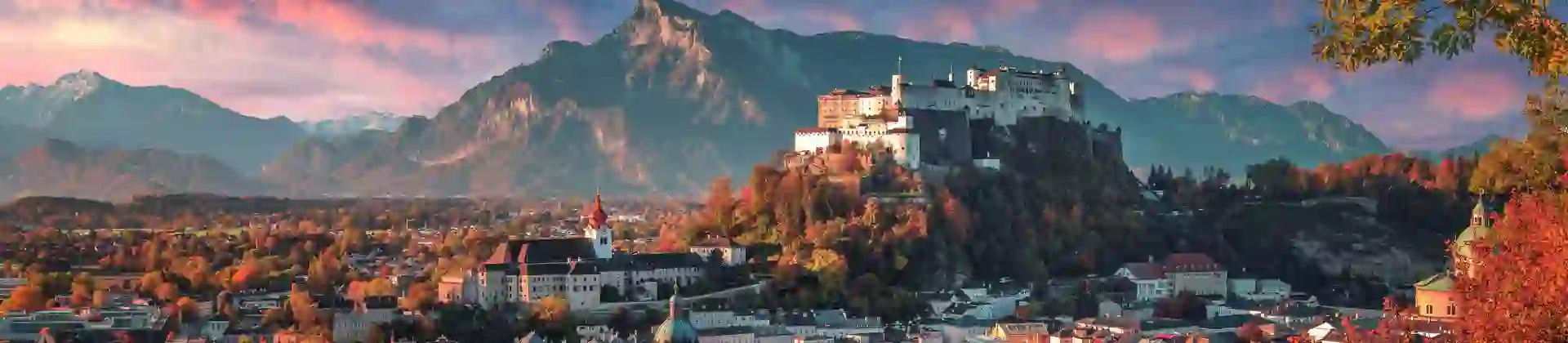 Wide angle shot of the city of Salzburg, showing the white and cream buildings, one with a turquoise turret and one further back with a red turret. Slightly to the right is a hill covered in green and orange trees with the fortress on the top. Mountains in the distance in front of blue and pink sky. In the right forefront, there is red and green tree leaves.