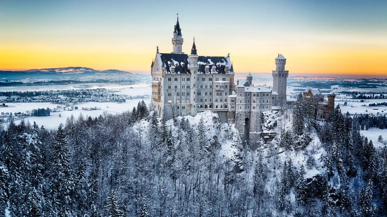 Wide angle shot Neuschwanstein Castle in the winter, showing its surrounding land covered in snow and a forest of fir trees in the forefront of the image. Behind the castle is a orange sky where the sun is setting