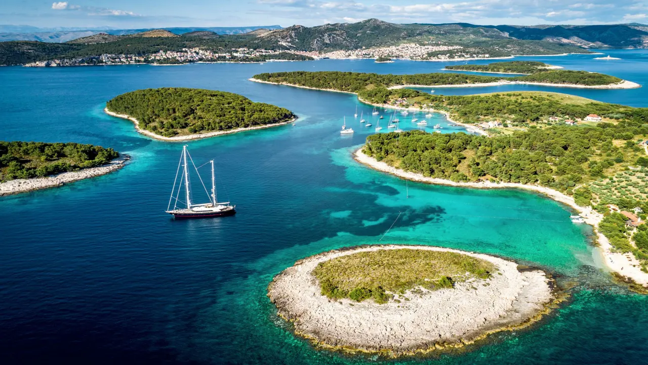 Aerial view of some small islands, surrounded by blue and turquoise water with a sailing boat near the forefront and a few more slightly further away. Mountains in the distance below a blue sky with some clouds.