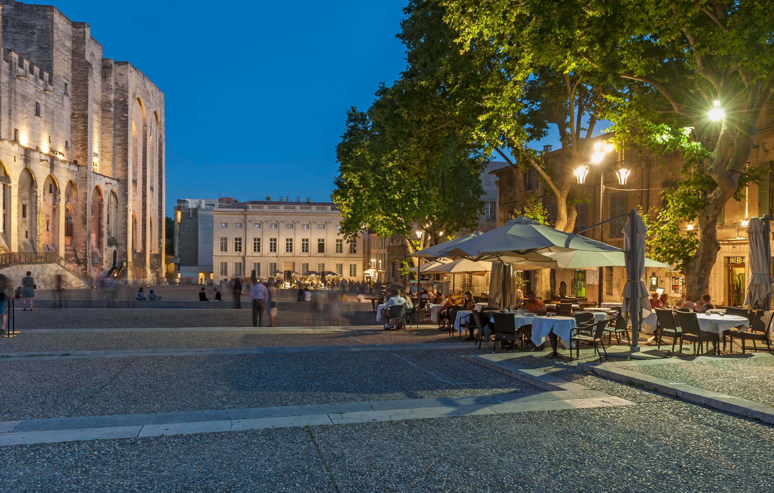 Outdoor seating outside The Palais Des Papes, that has white umbrellas, street lighting and trees above. A cream, wide manor is at the end of the road, and the edge of palace on the left.