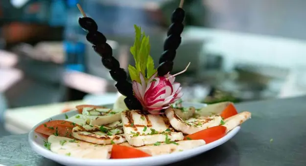 Close up of a dish of halloumi and tomatoes, topped with two skewers of olives, a radish cut into a flower shape, and herbs.