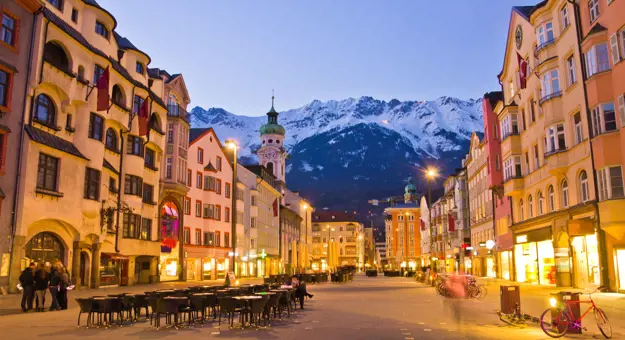 View of a wide road with shops and tall buildings either side. Tables and chairs are laid out in the road and bicycles are parked up on the right side. There is a clock tower which is taller than the rest of the buildings with a turquoise turret on the left, and snowy mountains can be seen at the end of the road, in front of light blue evening sky