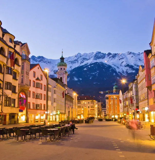 View of a wide road with shops and tall buildings either side. Tables and chairs are laid out in the road and bicycles are parked up on the right side. There is a clock tower which is taller than the rest of the buildings with a turquoise turret on the left, and snowy mountains can be seen at the end of the road, in front of light blue evening sky