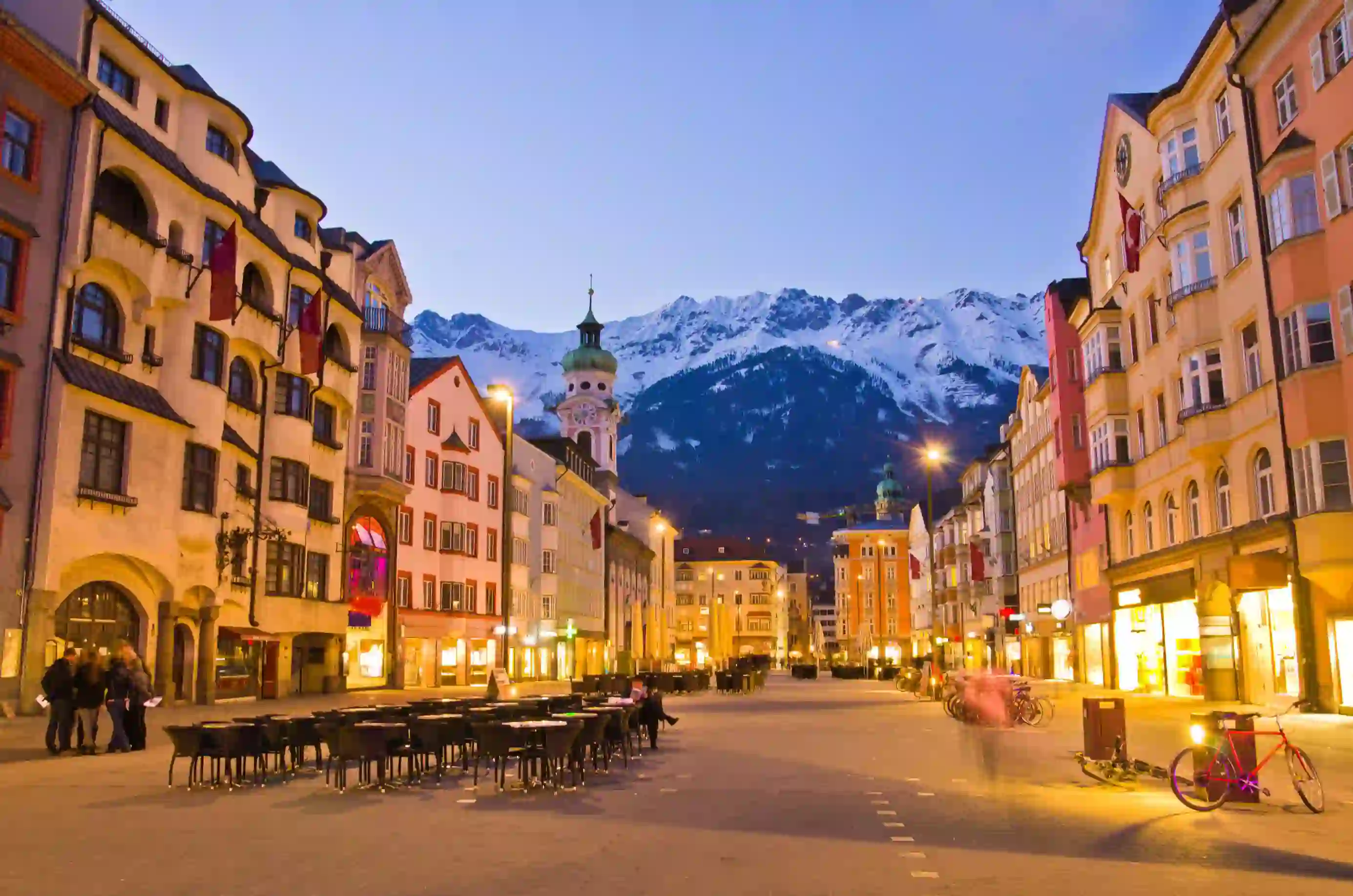 View of a wide road with shops and tall buildings either side. Tables and chairs are laid out in the road and bicycles are parked up on the right side. There is a clock tower which is taller than the rest of the buildings with a turquoise turret on the left, and snowy mountains can be seen at the end of the road, in front of light blue evening sky