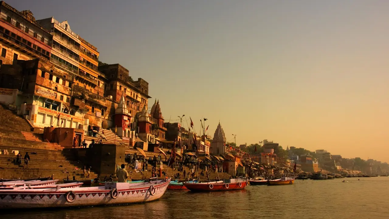 Boats anchored along the ghats of Varanasi at sunrise, with ancient sandstone buildings and temples glowing in golden light by the River Ganges
