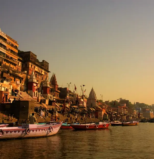 Boats anchored along the ghats of Varanasi at sunrise, with ancient sandstone buildings and temples glowing in golden light by the River Ganges