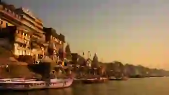 Boats anchored along the ghats of Varanasi at sunrise, with ancient sandstone buildings and temples glowing in golden light by the River Ganges