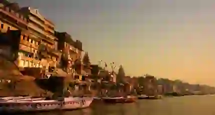 Boats anchored along the ghats of Varanasi at sunrise, with ancient sandstone buildings and temples glowing in golden light by the River Ganges