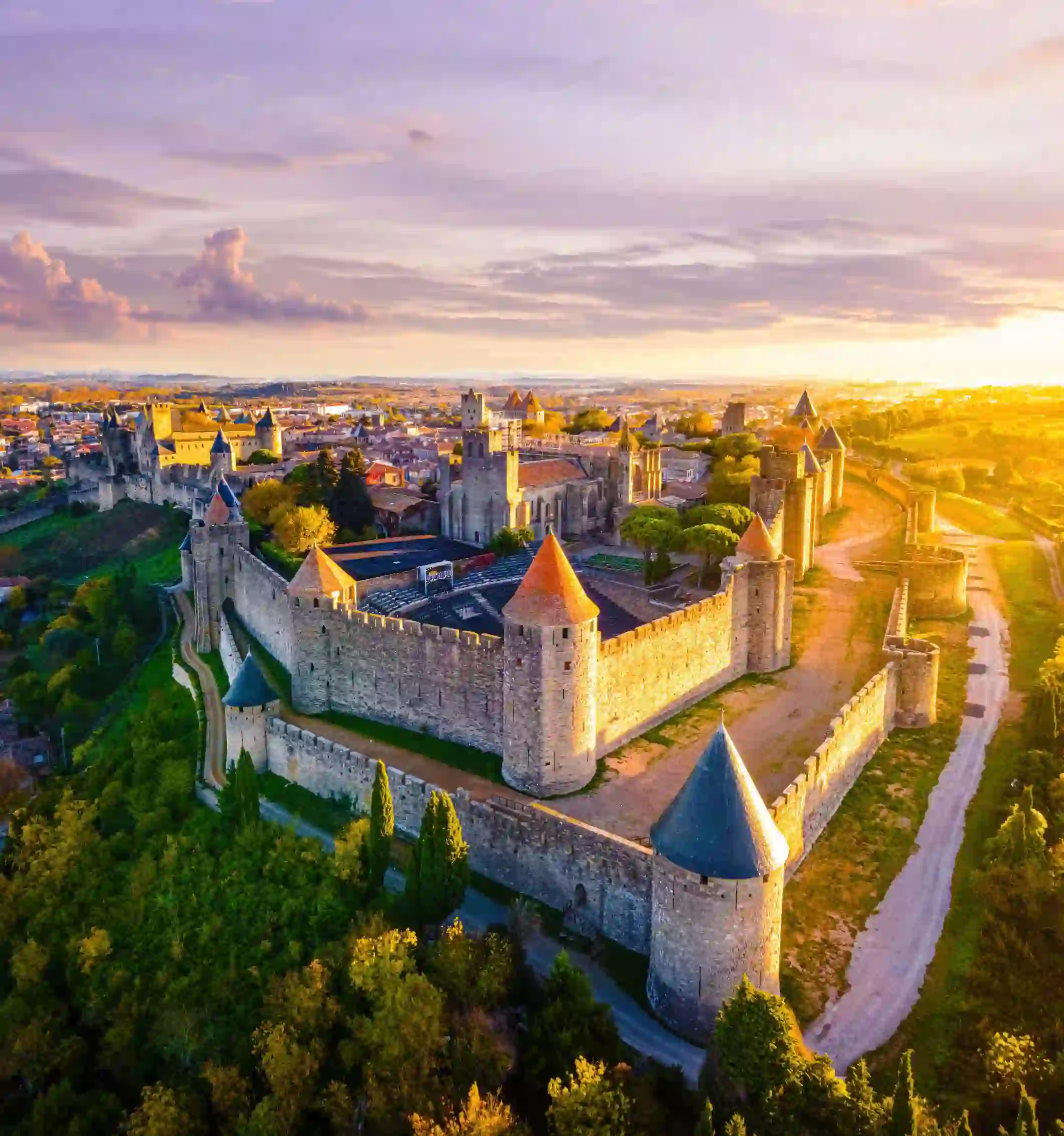 Bird's eye view of the Castle Of Carcassonne. Surrounded by trees and grass, and a pathway that loops around the castle. The castle has towers with red and blue turrets, and a distant view of a town can be seen behind it, under a grey/blue sky.