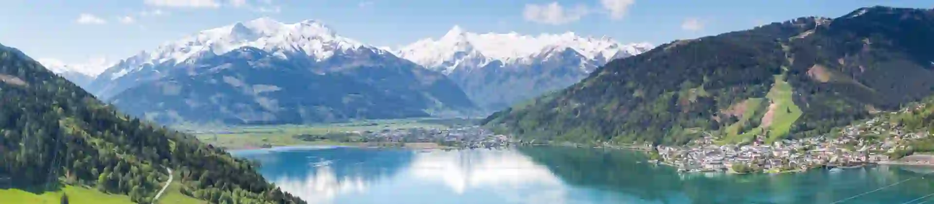 View of a large lake from a mountain, with houses and trees at the bottom. Another town can be seen on the other side of the lake to the right and centre of the image, and mountains with some snow on the top can be seen in the distance.