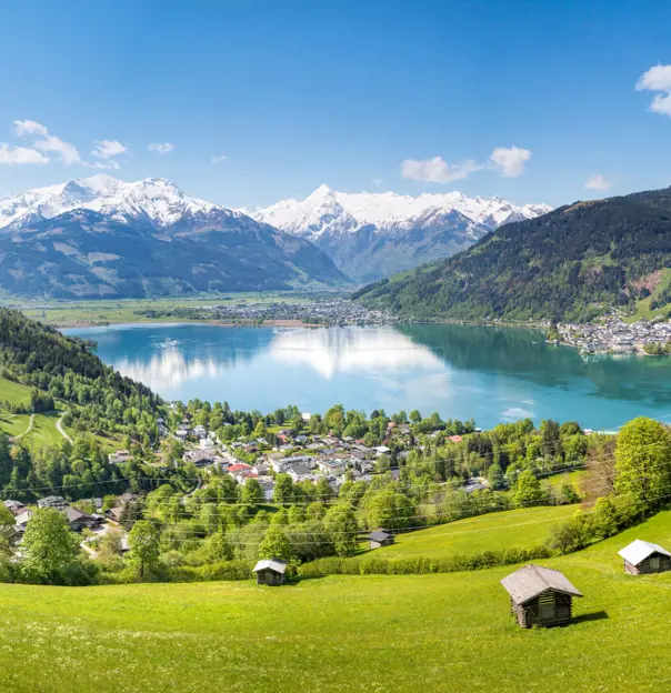 View of a large lake from a mountain, with houses and trees at the bottom. Another town can be seen on the other side of the lake to the right and centre of the image, and mountains with some snow on the top can be seen in the distance.