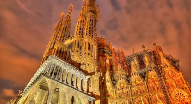 Night view of the illuminated Sagrada Família basilica in Barcelona, Spain, showcasing its intricate architecture and towering spires against a dramatic, cloudy sky