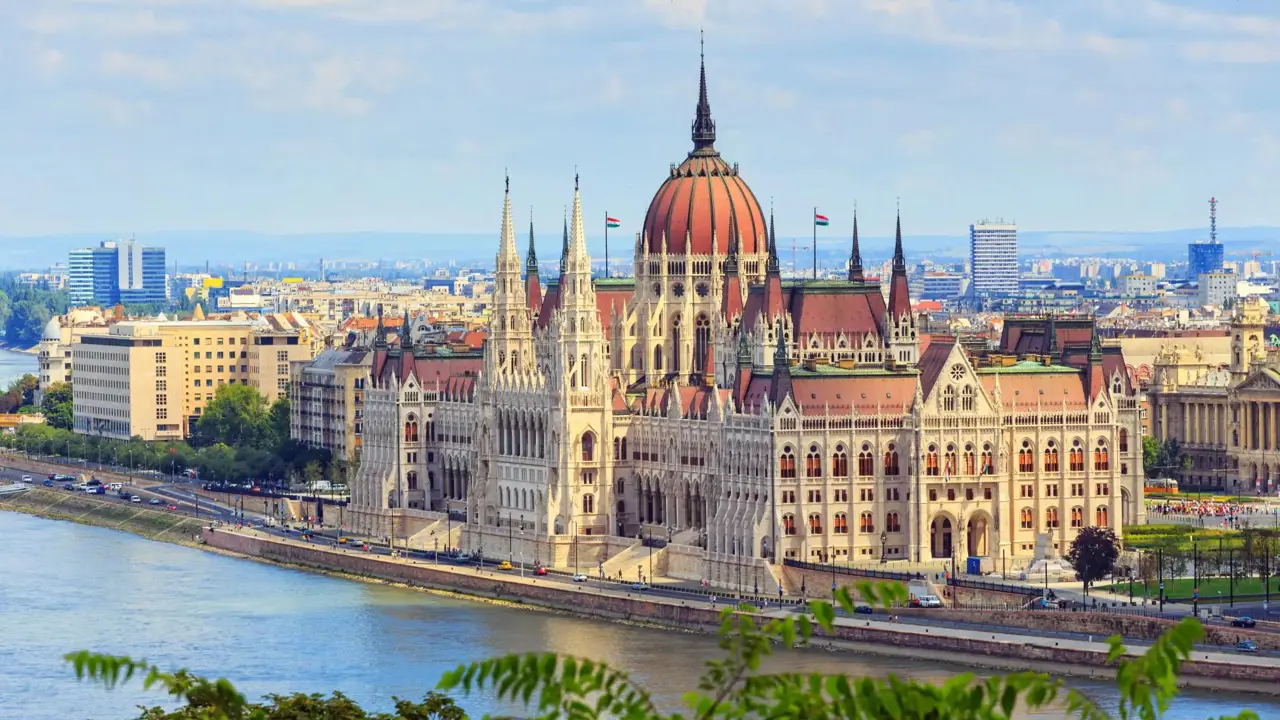 High angle shot of the Hungarian Parliament Building, with red roofs and spiky turrets and the Danube river in the left forefront