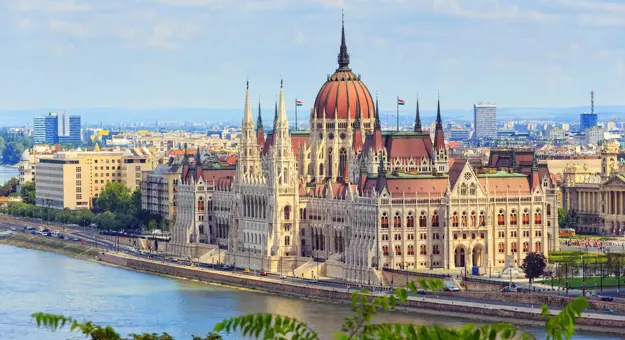 High angle shot of the Hungarian Parliament Building, with red roofs and spiky turrets and the Danube river in the left forefront
