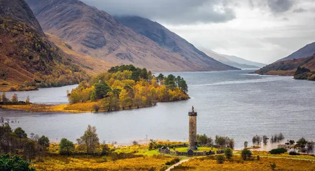 Glenfinnan Monument on a grassy hill at the head of Loch Shiel in the Scottish Highlands, with a lone statue of a Highlander atop the stone tower and dramatic mountains in the background