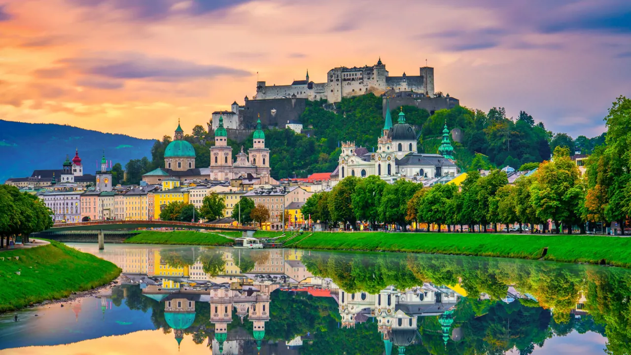 View of Salzburg from the river, showing houses along the river bed, churches and historical landmarks with turquoise turrets behind them, and the fortress on the top of the hill. There is a clear reflection of the city in the river at the forefront of the image, and the sky is a purple and orange sunset