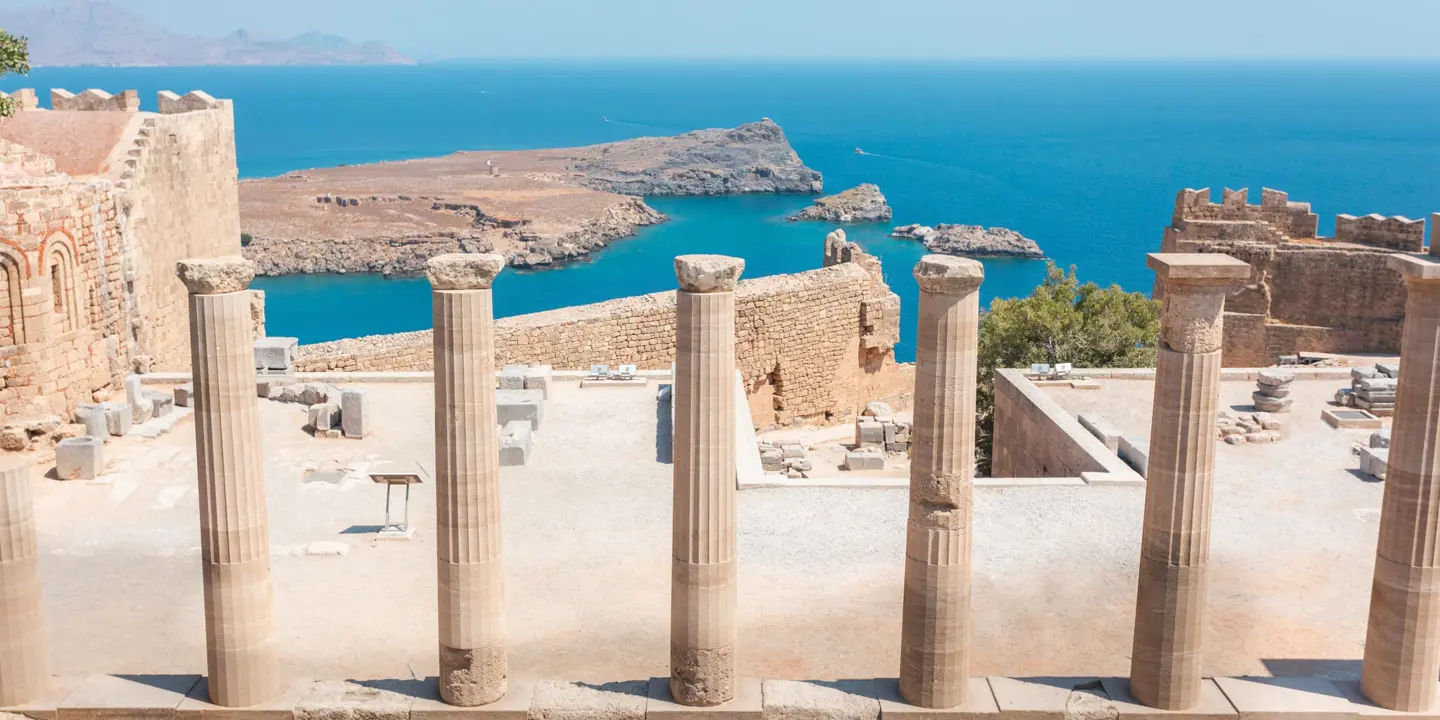 Stone pillars lined up in the forefront, with the ruins of Lindos city in front, and the view of the sea.