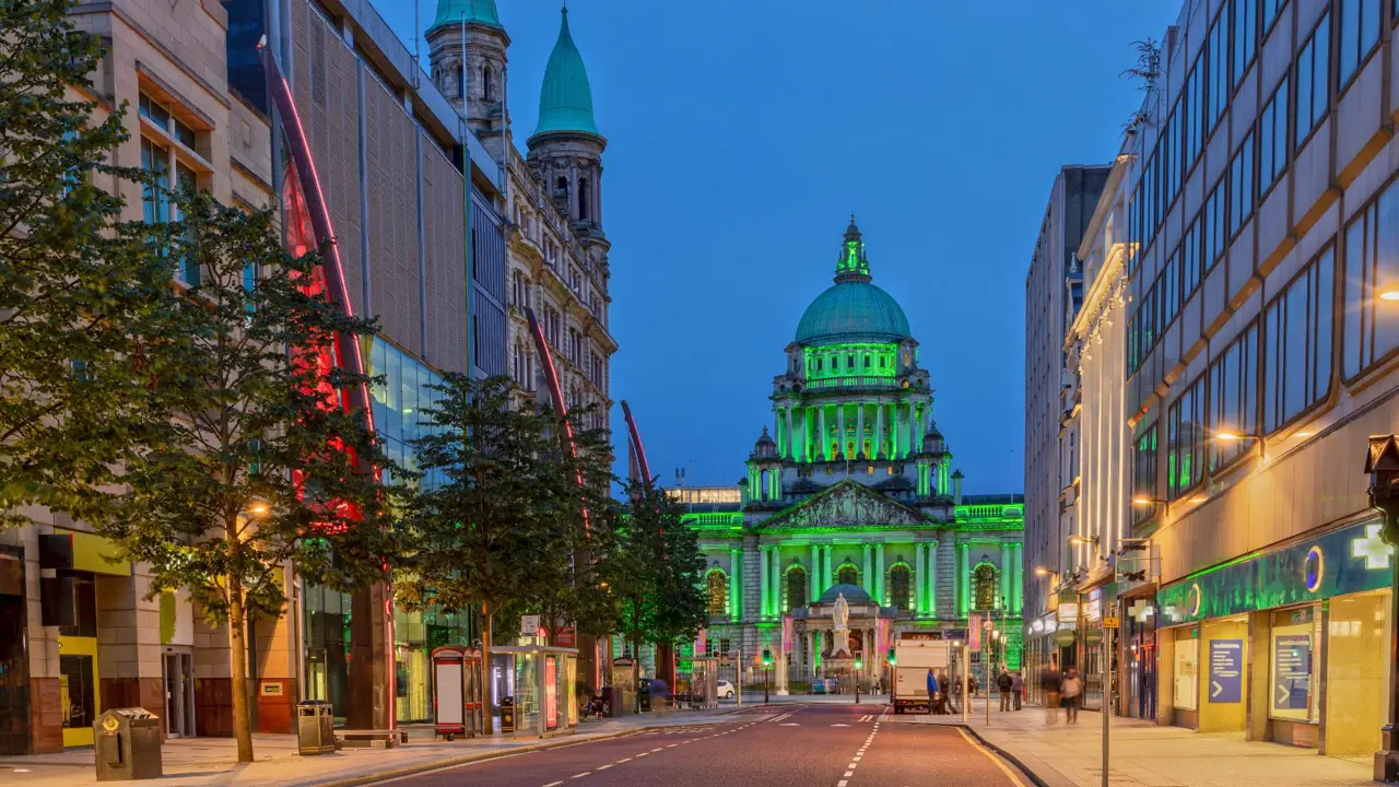 Evening view down Donegall Square with Belfast City Hall lit in green, framed by modern shops and historic buildings