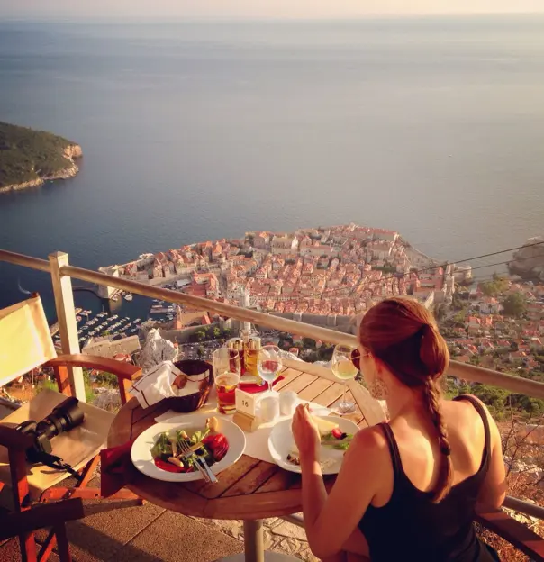 Back of a woman eating on a balcony overlooking Dubrovnik, the coastal town full of buildings with orange roofs on the seafront. On the table there is glasses of wine, a beer and bowls of salad