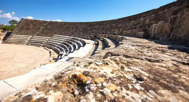 Low angle shot of the rows of seating in an ampitheatre, curving to the left, with a person sat on one of the rows on the far side. Above, a clear blue sky