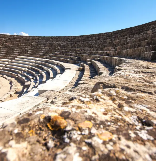 Low angle shot of the rows of seating in an ampitheatre, curving to the left, with a person sat on one of the rows on the far side. Above, a clear blue sky