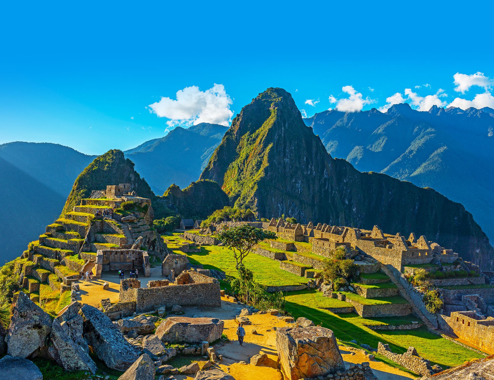 A panoramic view of Machu Picchu in Peru, showing ancient Incan ruins built into a green mountain ridge under a clear blue sky, with Huayna Picchu towering in the background