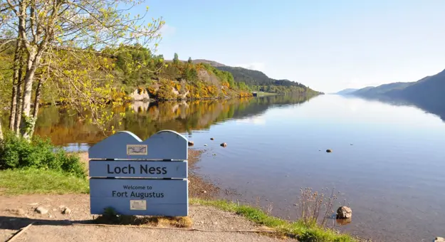 Calm waters of Loch Ness reflecting the surrounding hills under a clear blue sky, with a sign welcoming visitors to Fort Augustus on the shore
