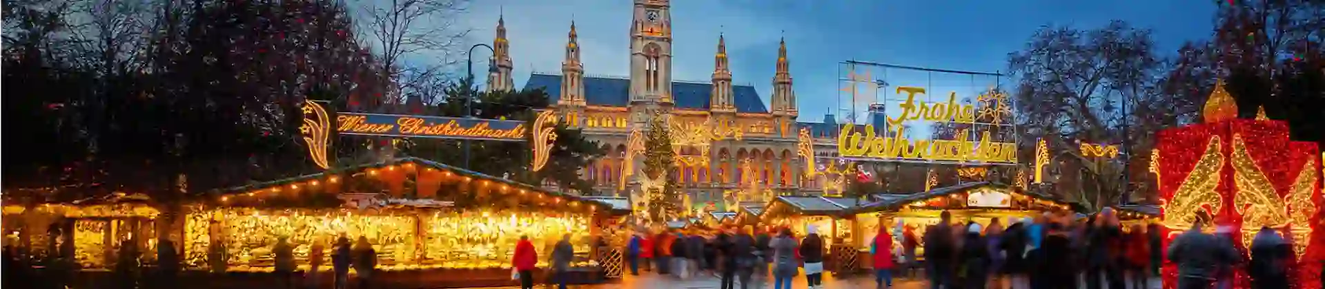Christmas market in front of Vienna’s City Hall at dusk, with festive lights, decorated stalls, and crowds enjoying the festive atmosphere. Signs read “Wiener Christkindlmarkt” and “Frohe Weihnachten"